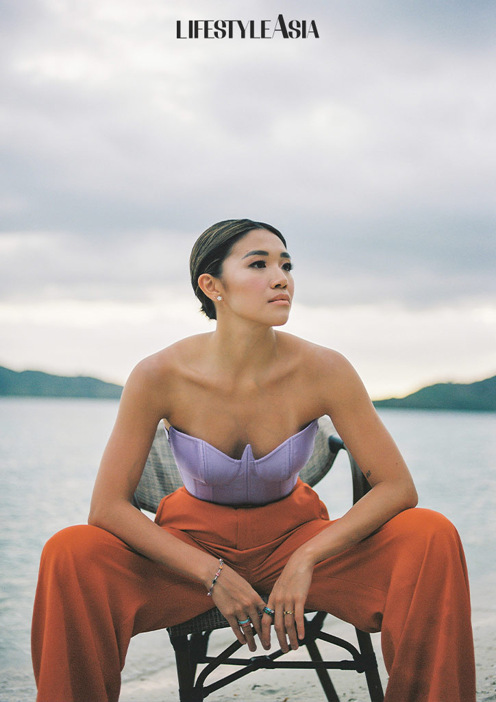 Seated on the beach while wearing Lilac bustier and burnt orange trousers by Rosenthal Tee, diamond earrings and multi-colored rings by Suki Jewelry, multi-colored tennis bracelet by Mei Diamond Jewelry.